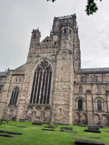 Durham Cathedral’s imposing stone exterior rises against a blue sky, showcasing Gothic-style arched windows, massive masonry walls, and historic architectural details. The weathered gray stonework and fortress-like design highlight the cathedral’s medieval character and centuries-old heritage.