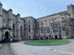 Historic stone building at Durham University featuring castle-like architecture with tall arched windows, crenellated walls, and round corner turrets topped with domed roofs. The gray masonry façade surrounds a grand arched entrance under a clear blue sky.
