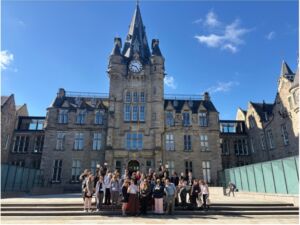 Group gathered outside the Edinburgh Futures Institute, standing before the landmark clock-towered stone building on a sunny day.