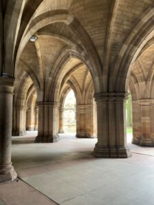 The Cloisters (Undercroft) at the University of Glasgow, with vaulted Gothic arches, stone columns, and light filtering through the open walkways.