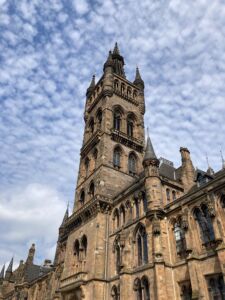 Low-angle view of the Gilbert Scott Building at the University of Glasgow, showing its ornate Gothic Revival stone façade and prominent central tower rising against a sky filled with scattered clouds. Arched windows, decorative stone carvings, and pointed turrets highlight the building’s historic architecture.