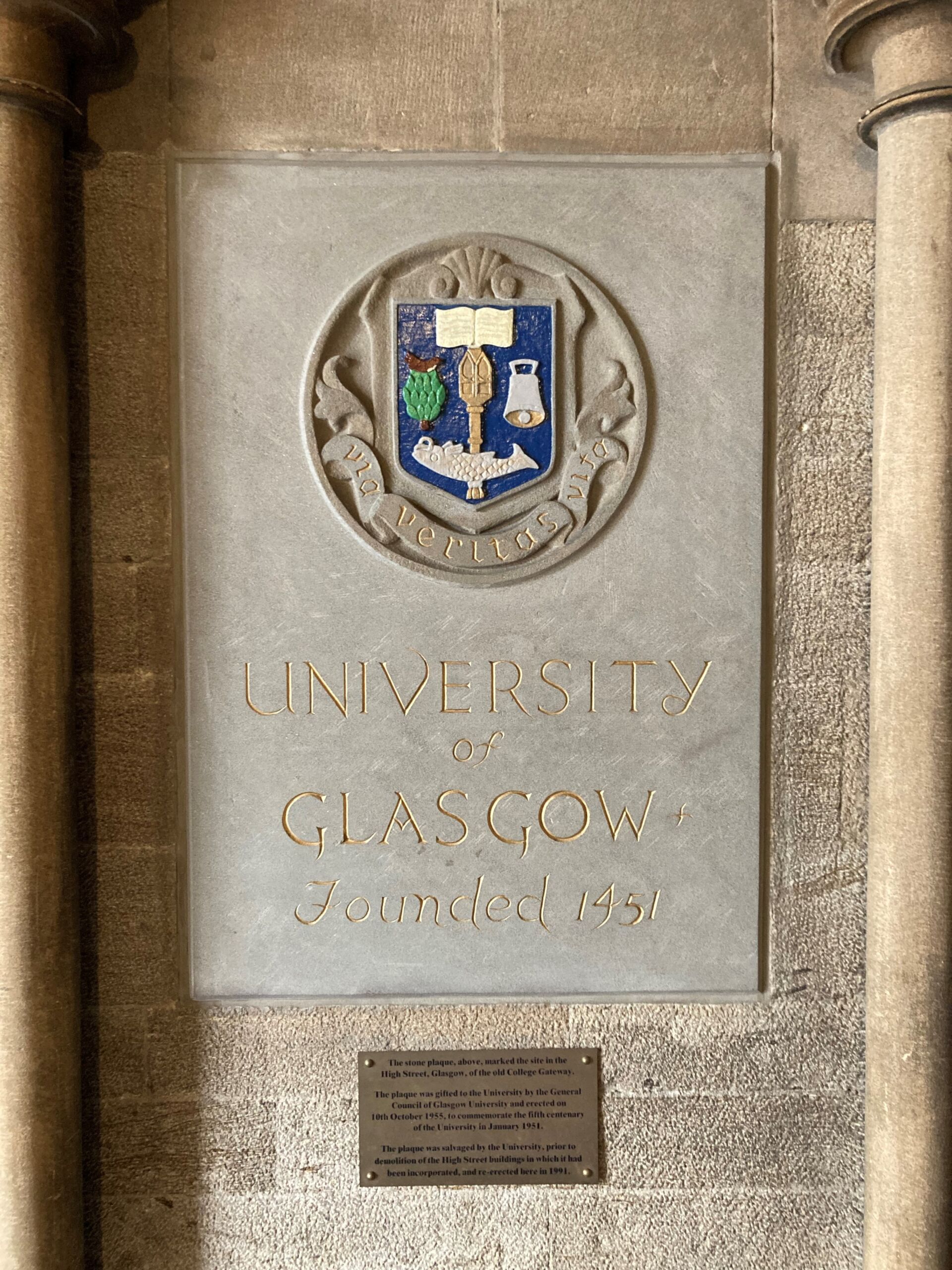 Stone plaque that reads University of Glasgow Founded 1451 with the university seal. Gold plate below plaque reads: The stone plaque, above, marked the He site in the High Street, Glasgow, of the old College Gateway. The plaque was gifted to the University by the General Council of Glasgow University and erected on the 10th October 1955, to commemorate the fifth centenary of the University in January 1951. The plaque was salvaged by the University, prior to demolition of the High Street buildings in which it had been incorporated, and re-erected here in 1991.