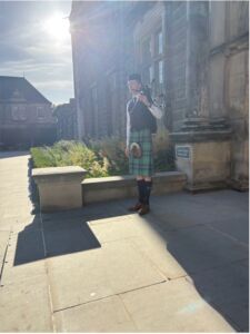 A bagpiper in traditional Scottish dress performs outside a historic University of St Andrews building on a sunny day.