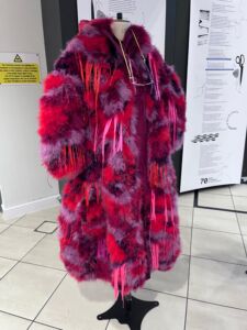 Example of student design piece. A long jacket with furry material that is multicolored red, pink, purple, and black. 