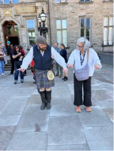 A participant learns traditional kilt folding from tour guide in Scottish dress in a stone courtyard.