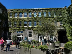 Exterior view of the Gates Cambridge building on a sunny day, showing a three-story brick façade covered in green ivy. A Gates Cambridge sign is visible near the main entrance.