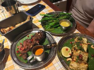 A metal platter lined with a green banana leaf holds several pieces of charred, spice-crusted grilled meat on the bone, garnished with scattered pomegranate seeds. A small metal jug of orange dipping sauce sits beside the meat. The platter is placed on a checkered tablecloth, with a basket of toasted flatbread on the right and additional dishes partially visible in the background.