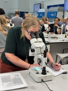 Woman with blonde hair and black glasses looking into microscope.