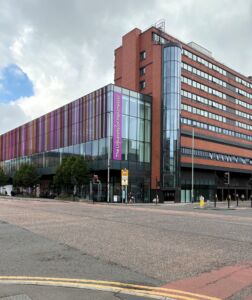 Modern multi-story University of Manchester building at a street corner featuring a red-brick tower and a long glass facade accented with vertical purple panels.