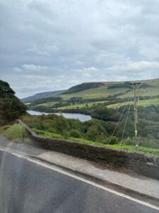 View from a coach window overlooking the Peak District countryside. A stone wall lines the roadside in the foreground. Beyond the wall, a winding reservoir sits among green fields, trees, and rolling hills.