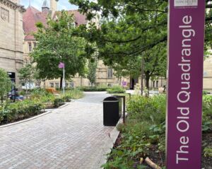 Brick-paved pedestrian walkway lined with trees, flowers, and landscaped garden beds on the University of Manchester campus. A tall purple sign in the foreground reads “The Old Quadrangle.” Stone academic buildings with red-tiled roofs are visible in the background.