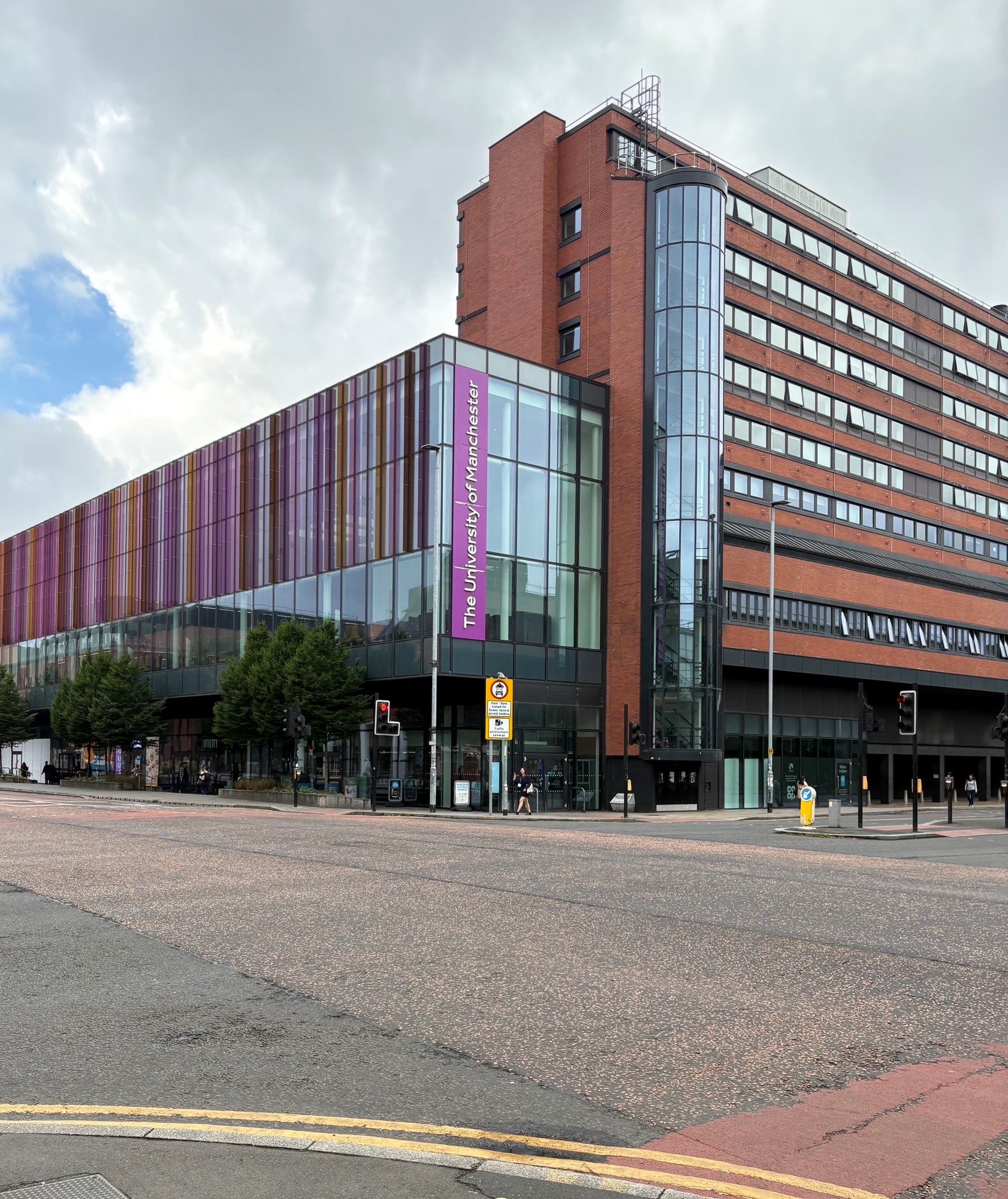 Modern multi-story University of Manchester building at a street corner featuring a red-brick tower and a long glass facade accented with vertical purple panels.