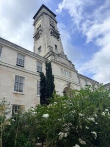 Trent Building at the University of Nottingham viewed from a low angle, featuring its tall stone clock tower rising against a partly cloudy blue sky. White stone walls and large windows frame the historic building, while flowering shrubs and greenery fill the foreground.
