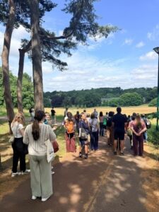 NAFAns participate in a walking tour of the University of Nottingham. Participants are gathered on a tree-lined campus path overlooking an open grassy field.