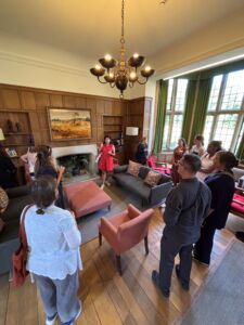 NAFAns gather in a room in the Rhodes house and appear to be listening to a current scholar. The room features large windows, two couches and chairs, and a fireplace with a plant inside of it. A landscape painting is hung above the fireplace.