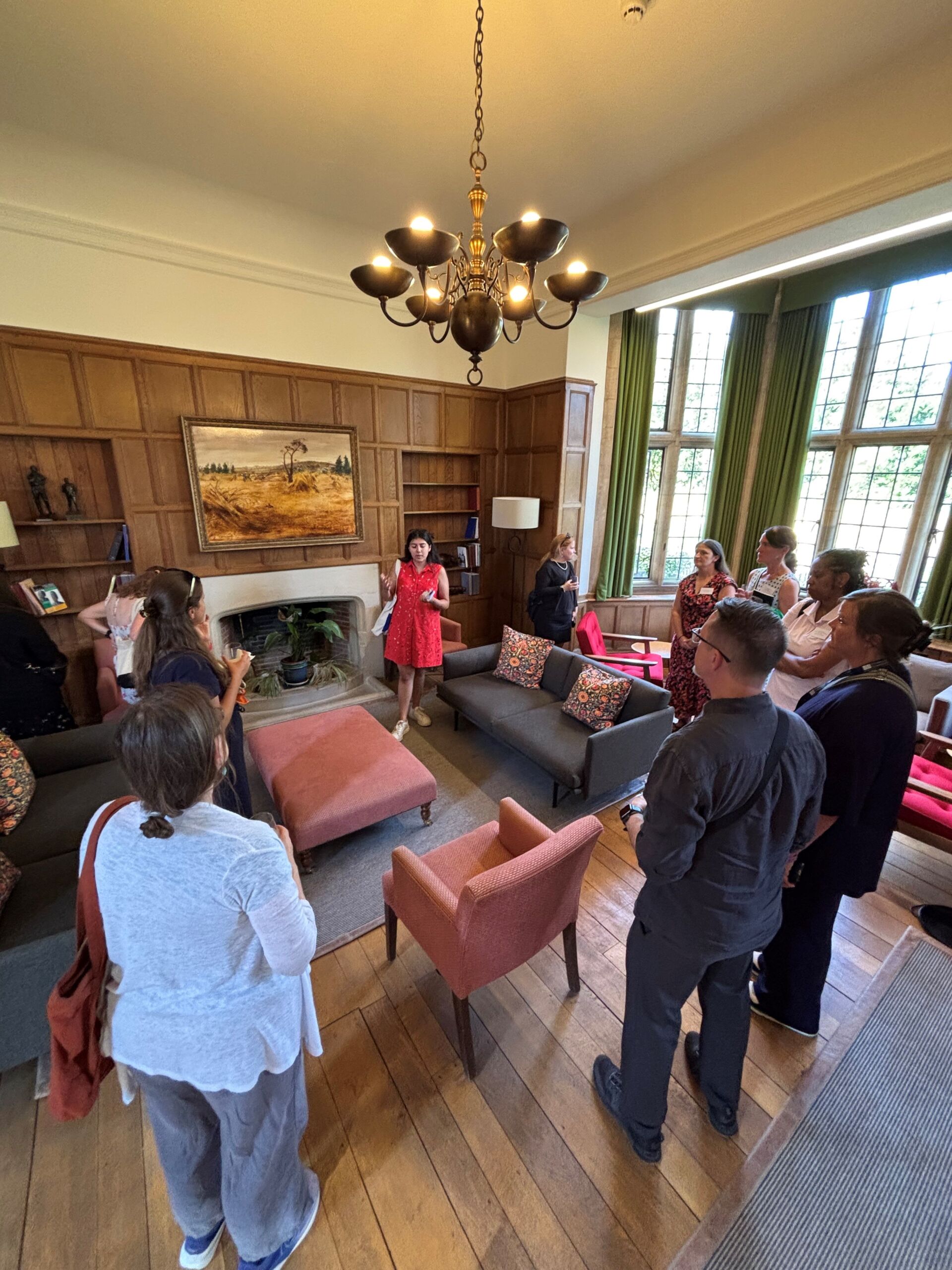 NAFAns gather in a room in the Rhodes house and appear to be listening to a current scholar. The room features large windows, two couches and chairs, and a fireplace with a plant inside of it. A landscape painting is hung above the fireplace.