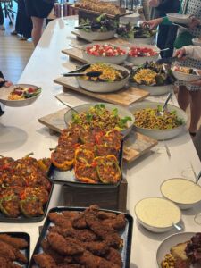 Table with bowls of food. There is fried chicken and stuffed peppers in the foreground, a chickpea salad and spicy coleslaw in the center, and at the top of the photo is a watermelon and feta salad.