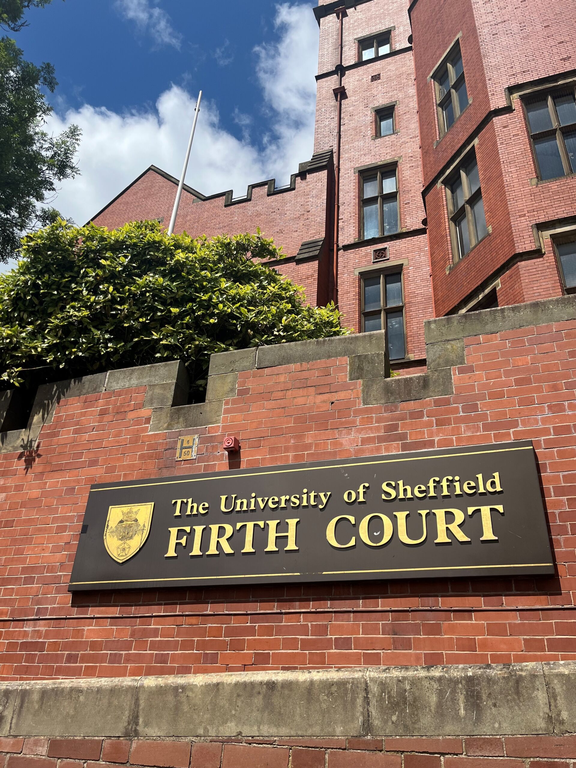 Low-angle view of a red-brick building with castle-like architectural details and tall rectangular windows. A large sign reads The University of Sheffield Firth Court