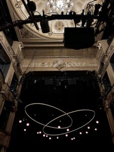 The stage set for Tom Stoppard’s Arcadia at the Duke of York’s Theatre, viewed from the auditorium. An ornate cream-and-gold Victorian interior with decorative plasterwork, balconies, and a chandelier frames the stage. Suspended above the darkened set are lighting rigs, speakers, and technical equipment. At center stage, a contemporary lighting installation of two intersecting glowing loops hangs against a black backdrop, surrounded by small pink illuminated spheres.
