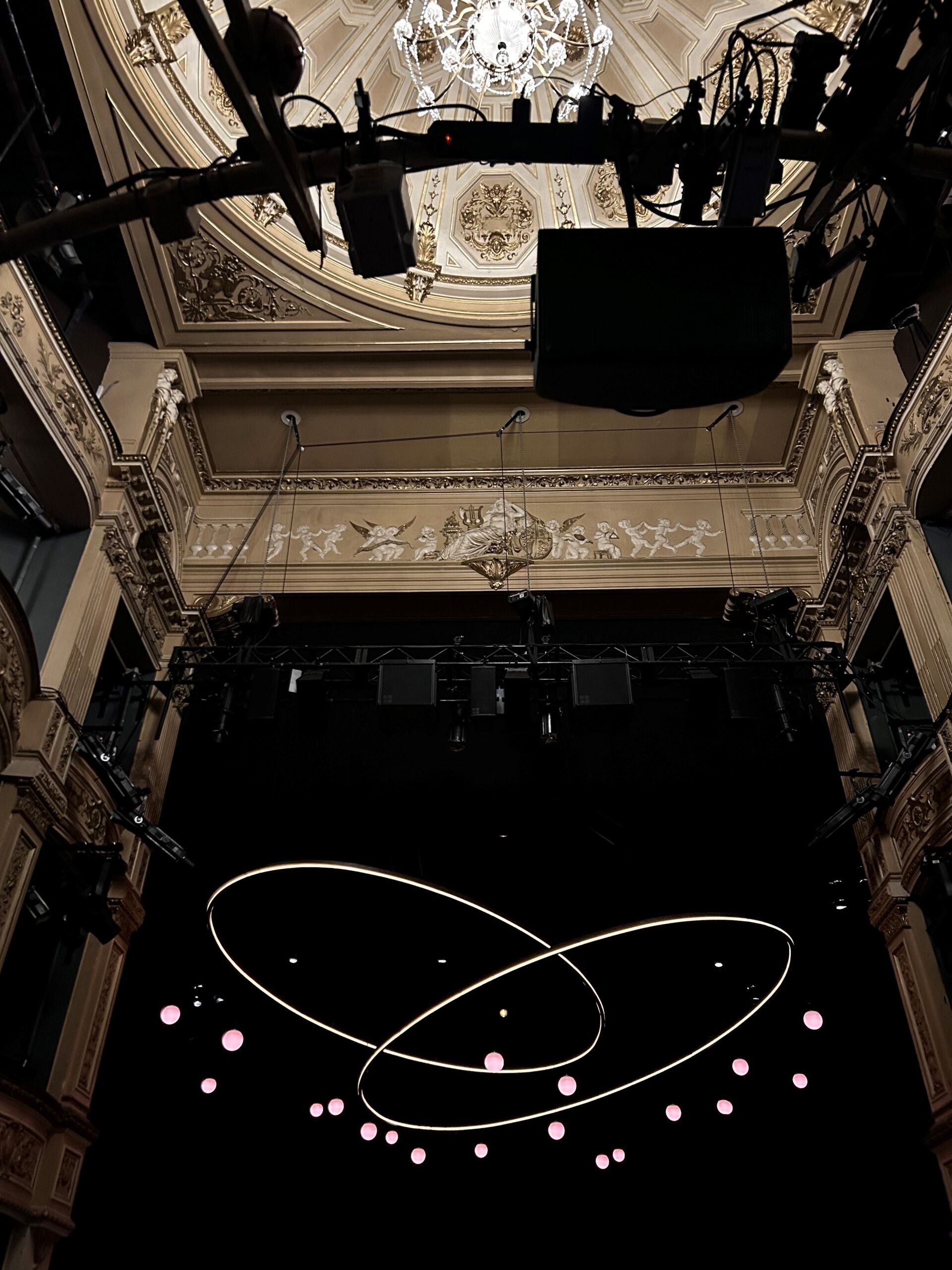 The stage set for Tom Stoppard’s Arcadia at the Duke of York’s Theatre, viewed from the auditorium. An ornate cream-and-gold Victorian interior with decorative plasterwork, balconies, and a chandelier frames the stage. Suspended above the darkened set are lighting rigs, speakers, and technical equipment. At center stage, a contemporary lighting installation of two intersecting glowing loops hangs against a black backdrop, surrounded by small pink illuminated spheres.