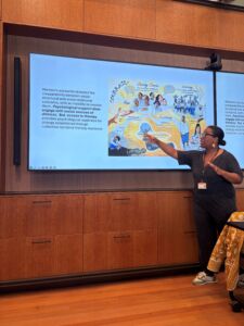 Professor Rochelle Burgess, a black woman with hair in a bun motions to a slide which highlights the Courage+ program in South Africa