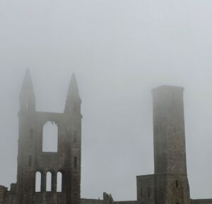 Stone ruins of the St Andrews Cathedral emerge from dense gray fog. On the left, a tall façade with two pointed towers frames a large arched opening and three narrow arched windows below. To the right stands a separate square stone tower.