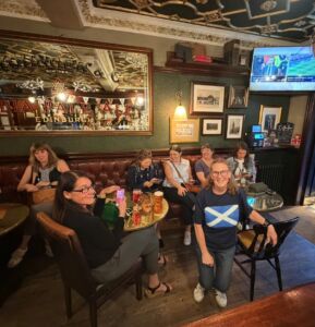 Several people are gathered around small tables in a pub. A large mirrored sign on the wall reads “India Pale Ale Edinburgh.” One person kneels in the foreground wearing a navy T‑shirt with a Scottish flag. In the background a television screen shows the World Cup.