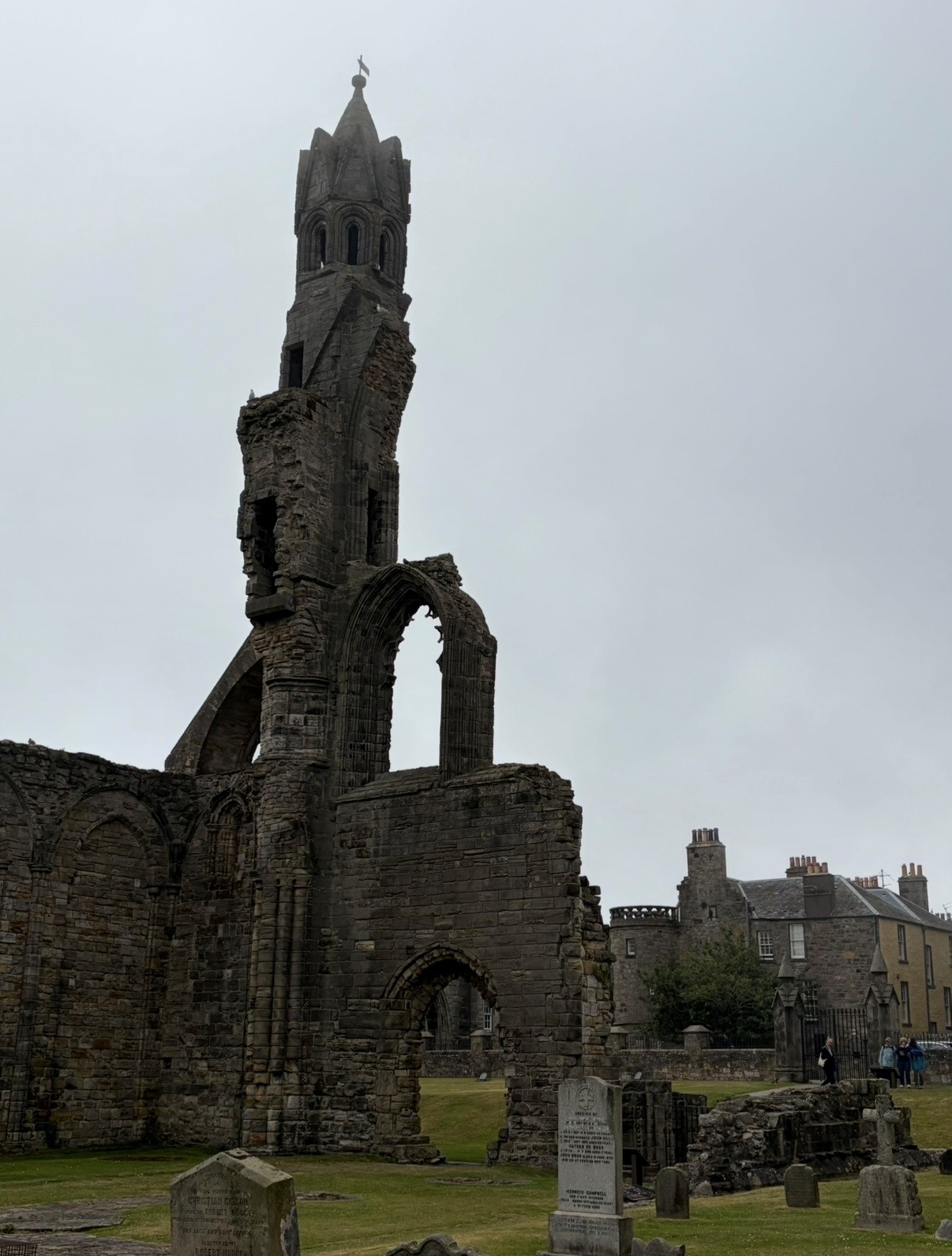 Ruins of St Andrews Cathedral stand beneath a gray overcast sky. A tall, partially intact stone tower with Gothic architectural details stands above the remaining walls and arches of the medieval cathedral. In the foreground, gravestones are scattered across a grassy churchyard.
